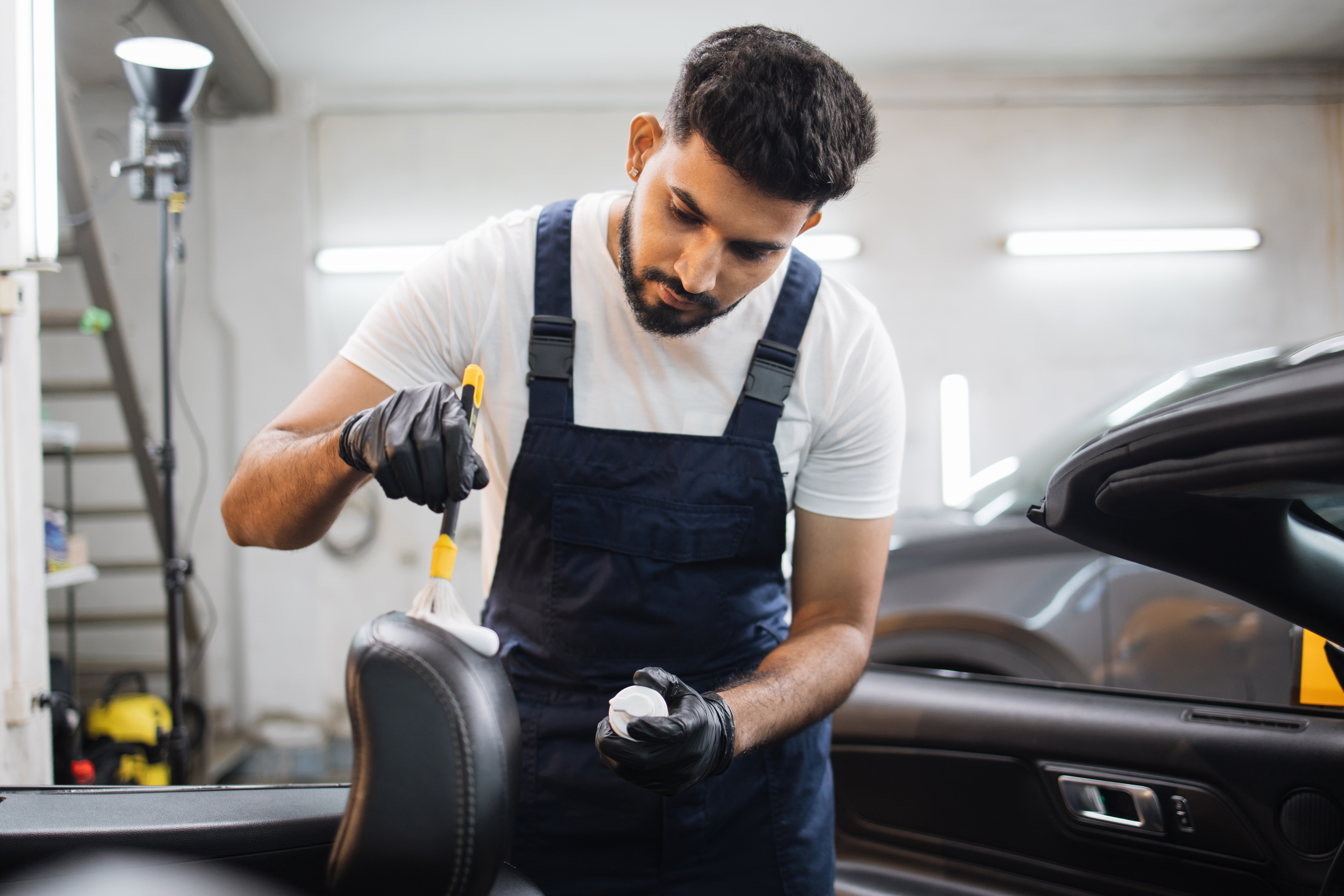 Young man in overalls and protective gloves, cleaning car seat with soft brush and foam.